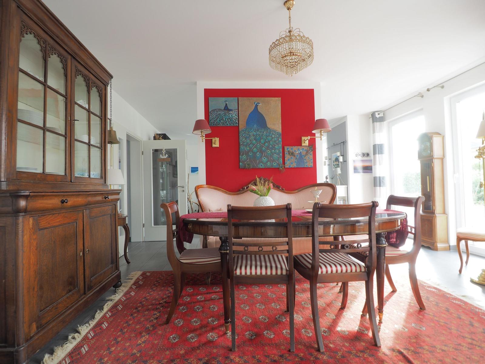 Esszimmer mit Holztisch, Stühlen und rotem Teppich. Wandbild mit Pfauen. Kronleuchter und antiker Schrank.