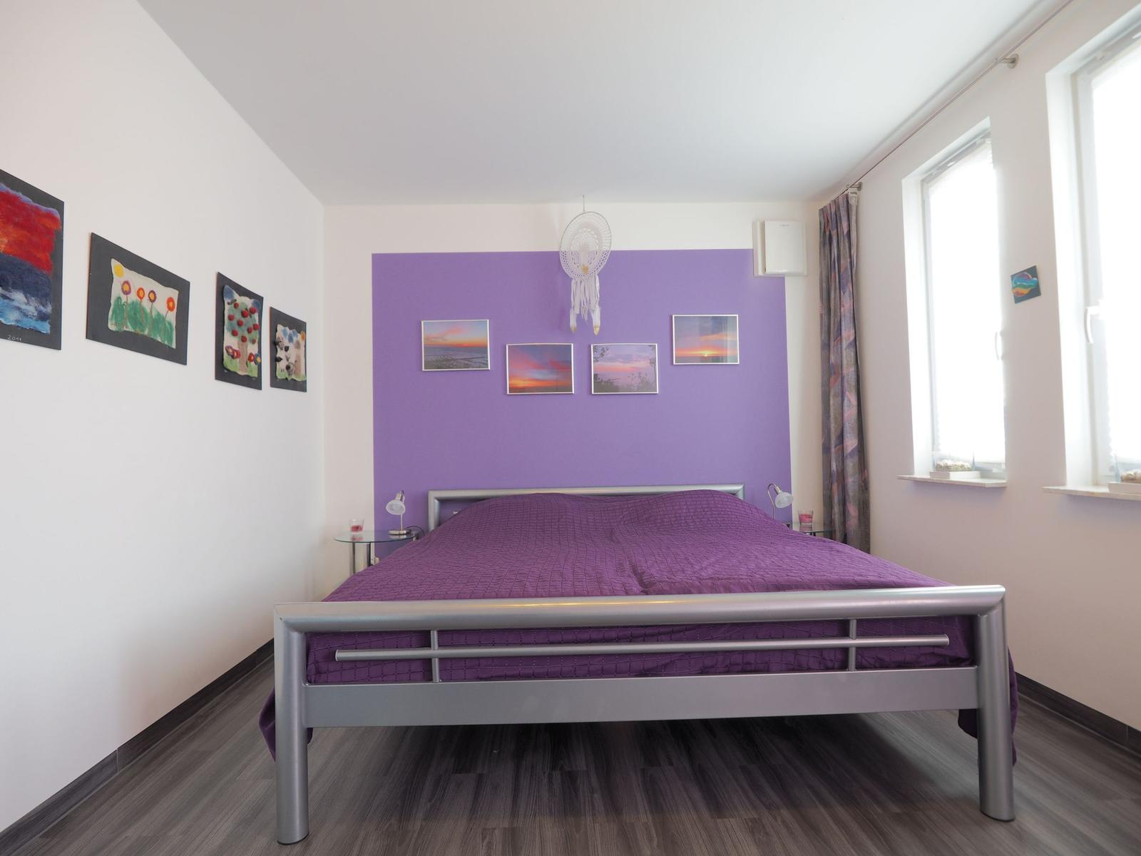 Schlafzimmer mit lila Wand, Bett und Fenstern.