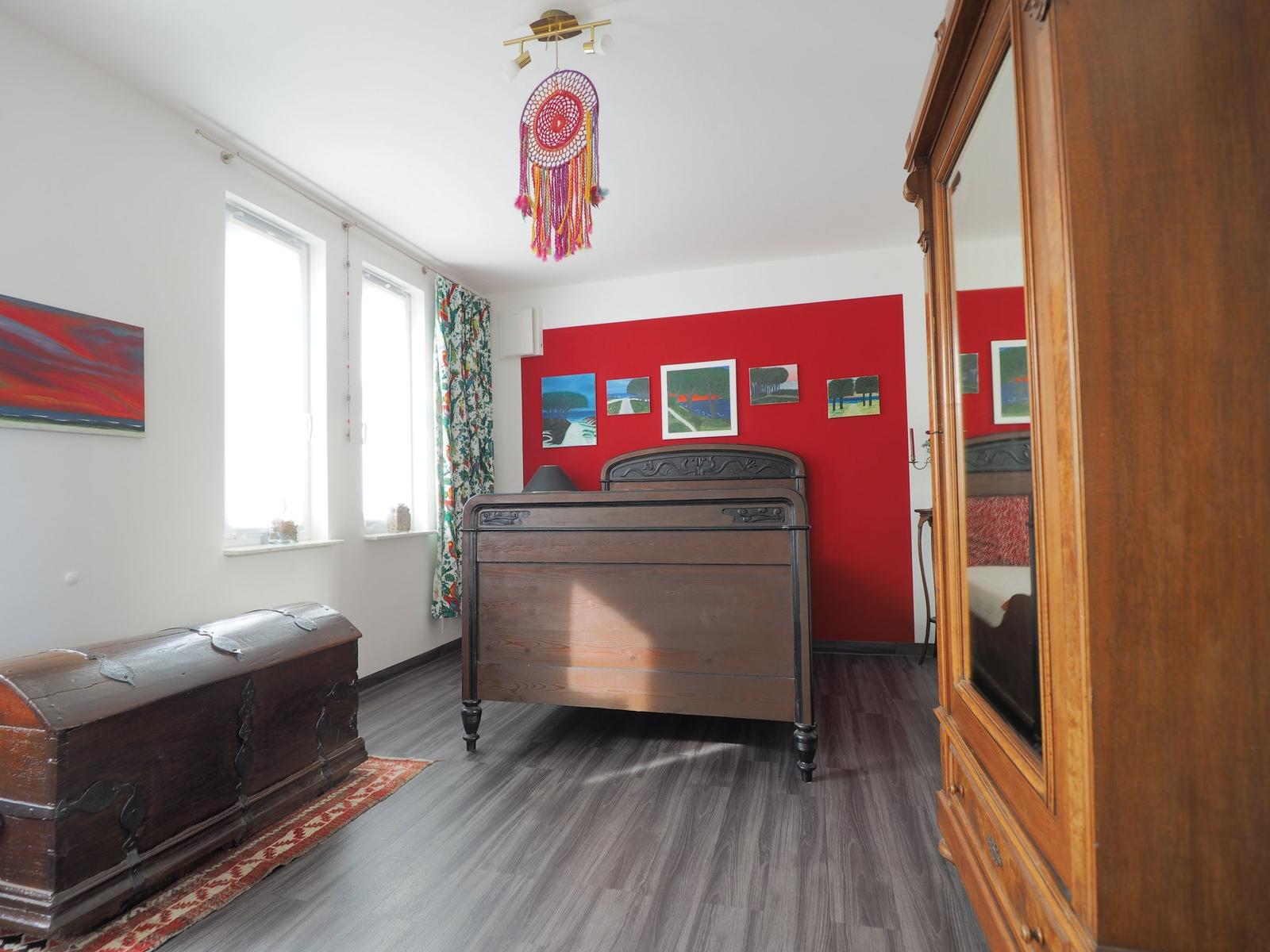 Schlafzimmer mit Holzbett, roter Wand, Spiegelschrank und Fenstern.