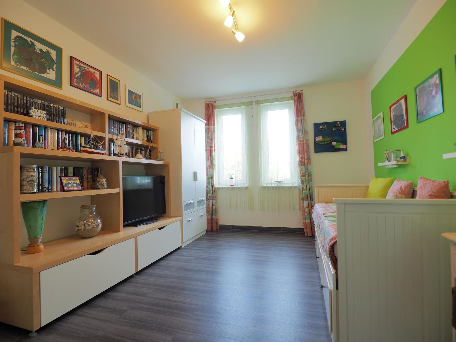 Ein Schlafzimmer mit Bett, Bücherregal, TV und Fenster mit Vorhängen.