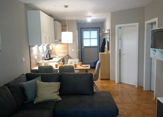 Wohn- und Essbereich mit Küchenzeile, Sofa und Tisch. Hintergrund: Tür und Kleiderständer.