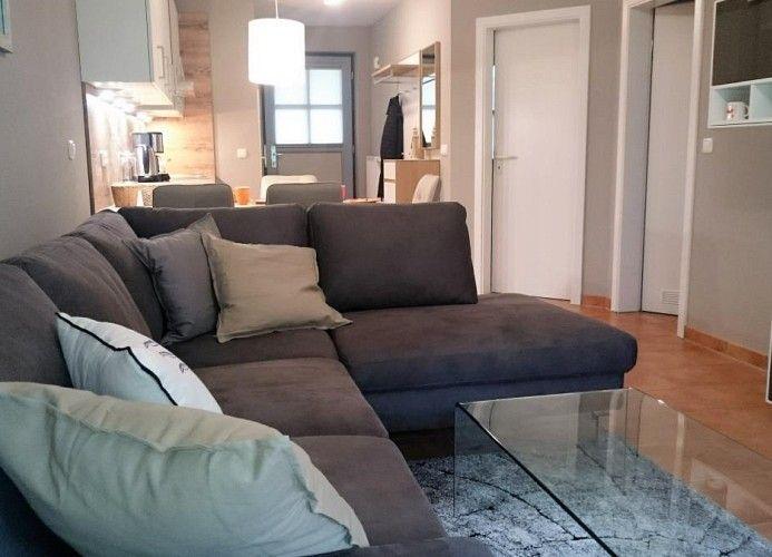 Ein Wohnzimmer mit grauem Sofa, Glas-Tisch und Küchenbereich im Hintergrund.