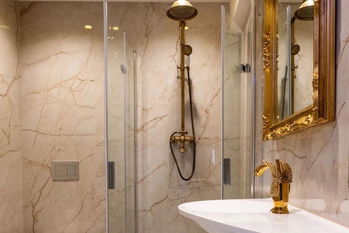 Bad mit Dusche, Marmorwand und goldfarbenem Waschbeckenhahn.