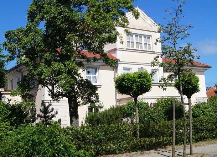 Weißes Haus mit rotem Dach, umgeben von Bäumen und Sträuchern unter blauem Himmel.