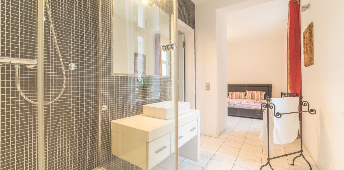 Badezimmer mit Dusche und Waschbecken. Im Hintergrund Schlafzimmer mit Bett.