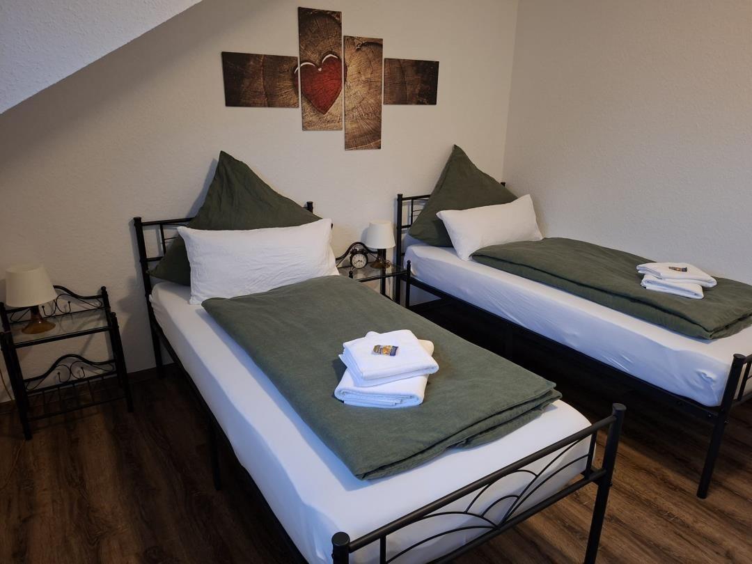 Zwei Betten mit grünen Decken und weißen Kissen. Wandbild mit Herz. Holzboden.