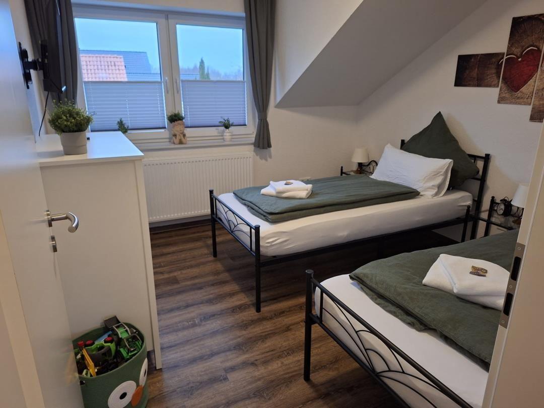 Zwei Betten mit grünen Decken, Fenster mit Jalousien und Holzboden.