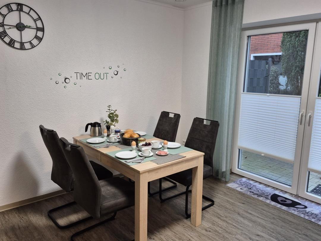 Esszimmer mit Tisch, Stühlen und Fenster. Wanduhr und Dekoration sichtbar.