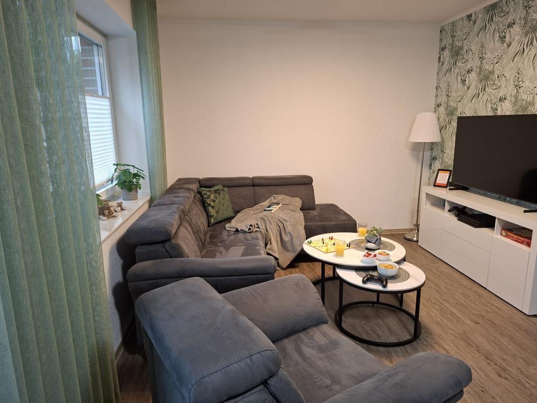 Wohnzimmer mit Sofa, Couchtisch und Fernseher. Grüne Vorhänge und Tapete.