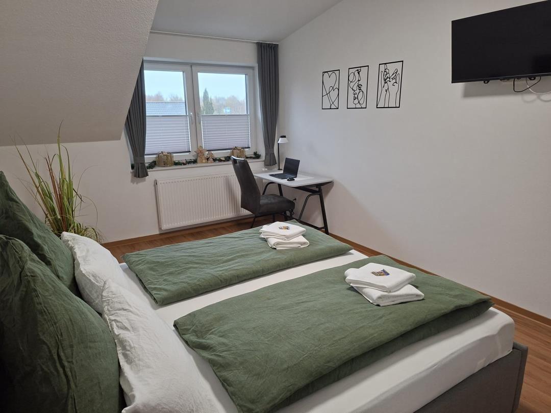 Schlafzimmer mit Doppelbett, Schreibtisch und TV. Fenster mit Jalousien und Wandbildern.