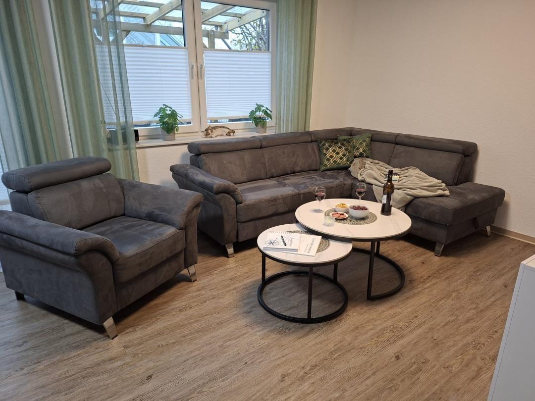 Wohnzimmer mit grauem Sofa, Sitzgelegenheit und Tischen. Fenster mit Jalousien und Pflanzen.