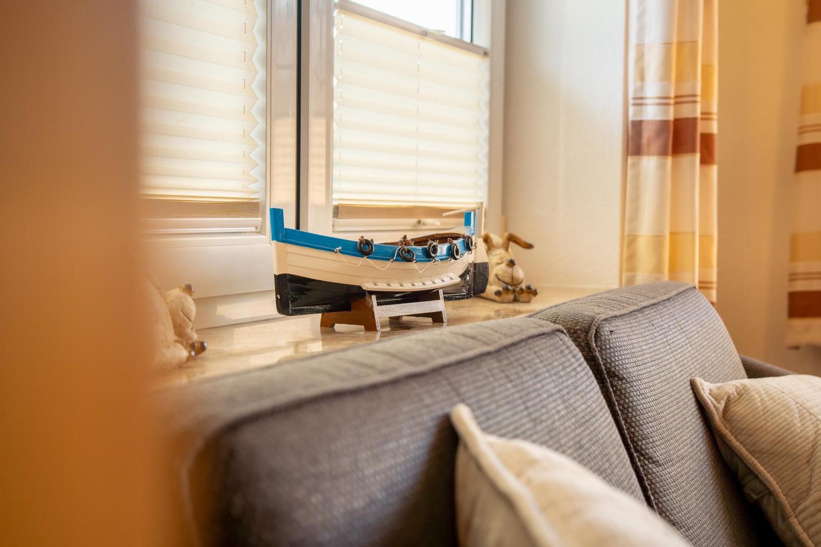 Ein grauer Sofa mit Kissen. Auf dem Fensterbrett steht ein kleines Boot und eine Figur.