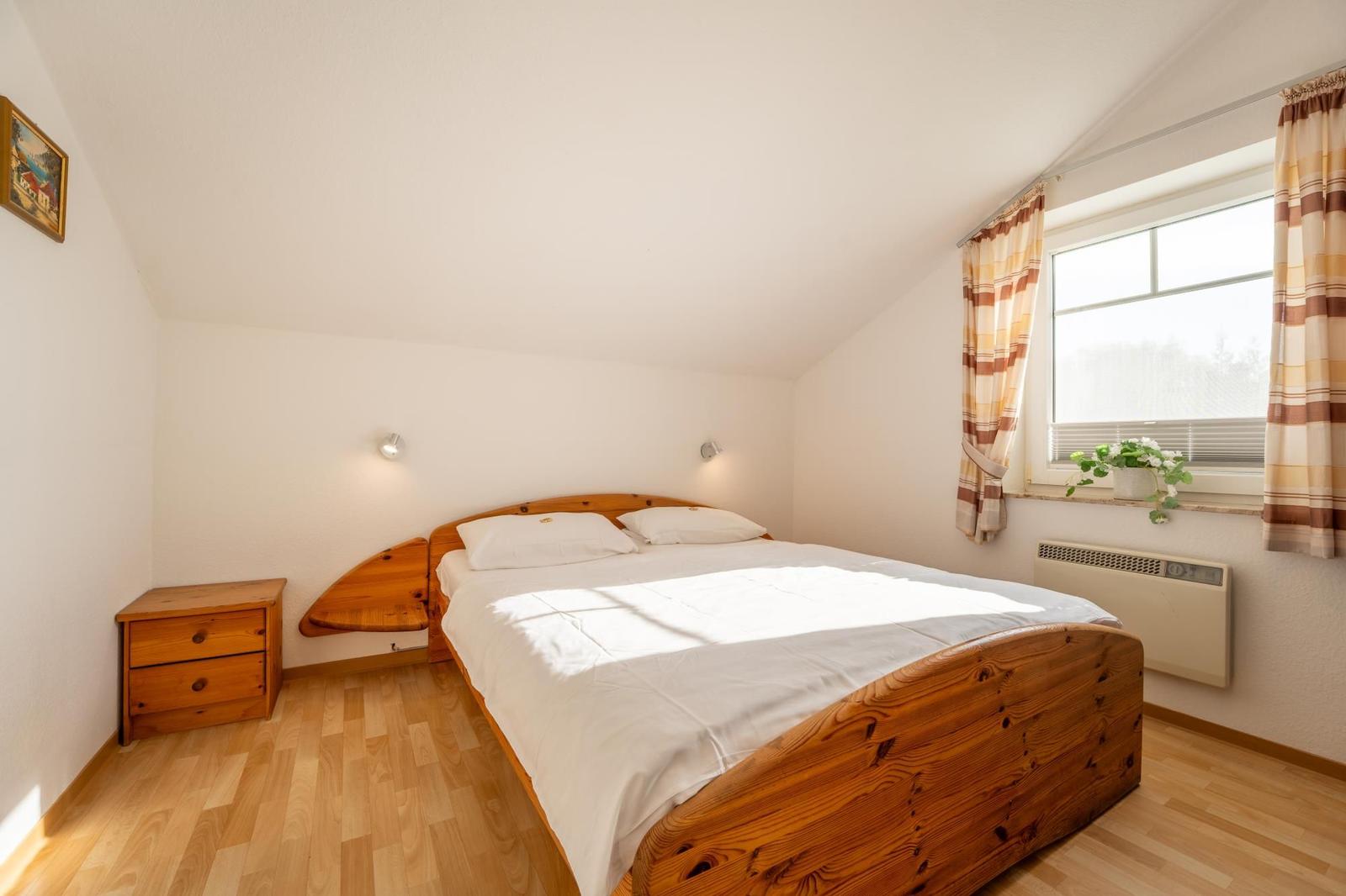 Schlafzimmer mit Holzbett, Nachttisch und Fenster mit Vorhängen.