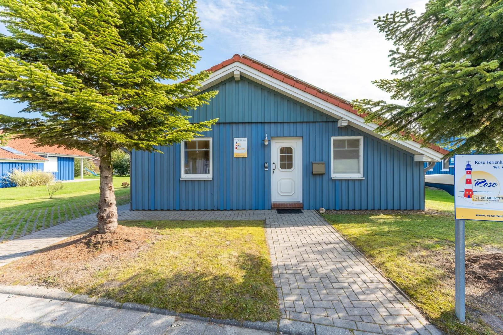 Blau gestrichenes Ferienhaus mit weißen Fenstern und Tür. Vor dem Haus liegt ein gepflasterter Weg.