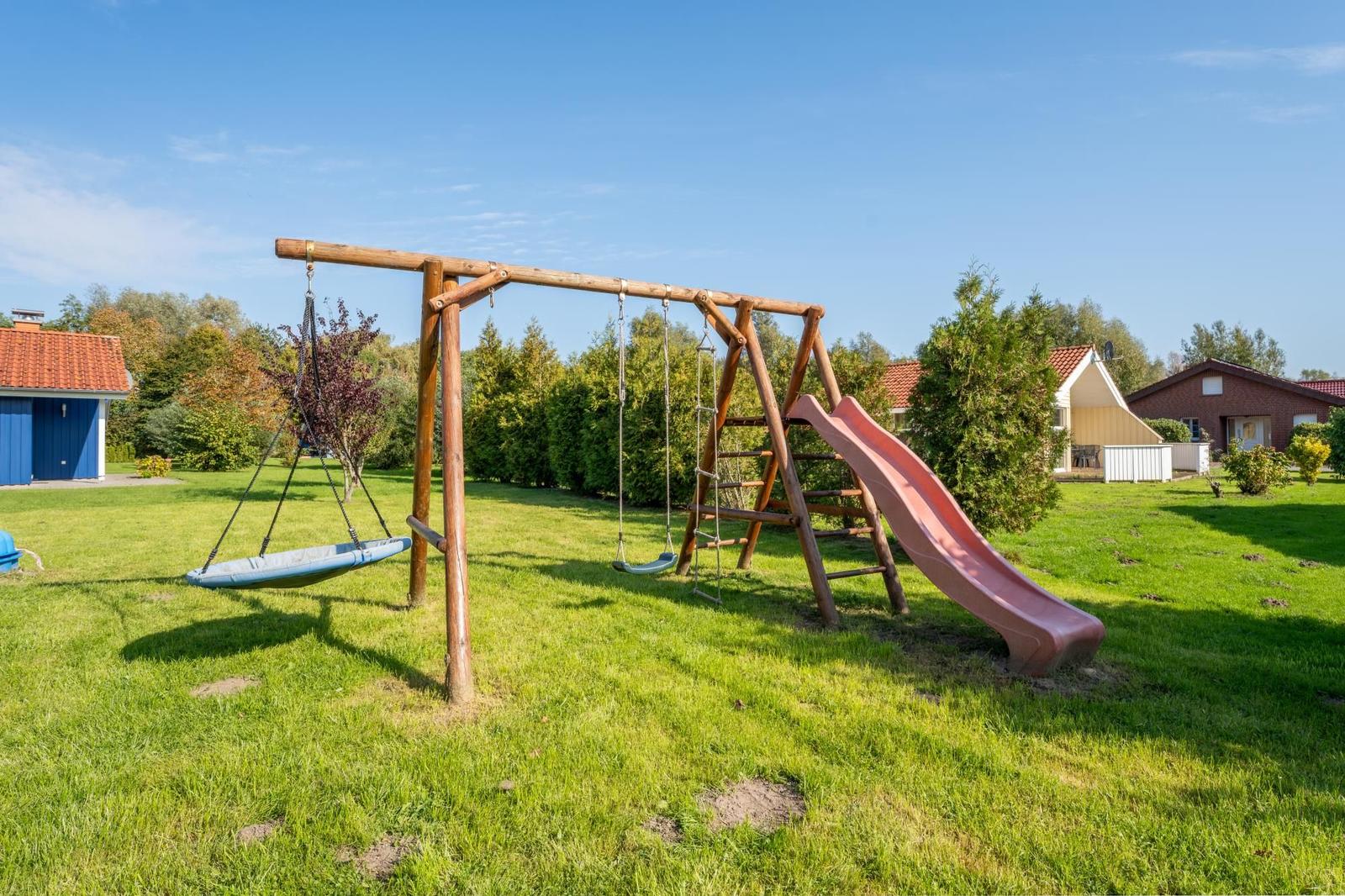 Holzspielplatz mit Rutsche, Schaukel und Klettergerüst auf grünem Rasen.