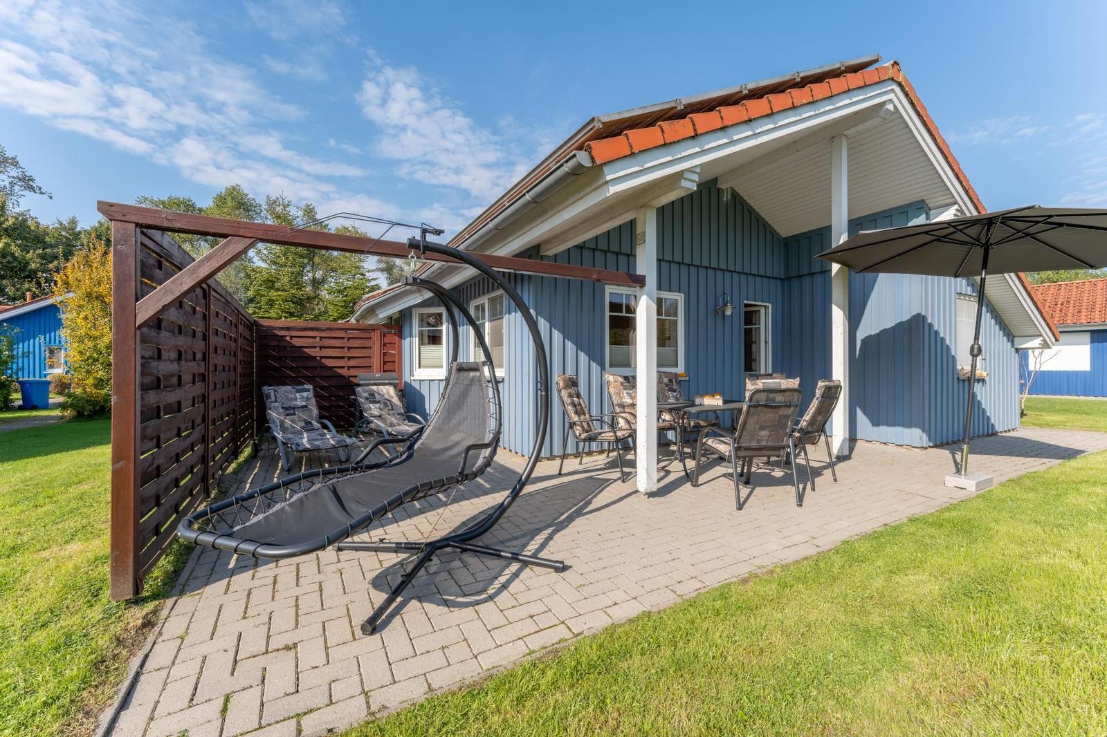 Terrasse mit Sitzgelegenheiten und Sonnenschutz vor einem blauen Haus.