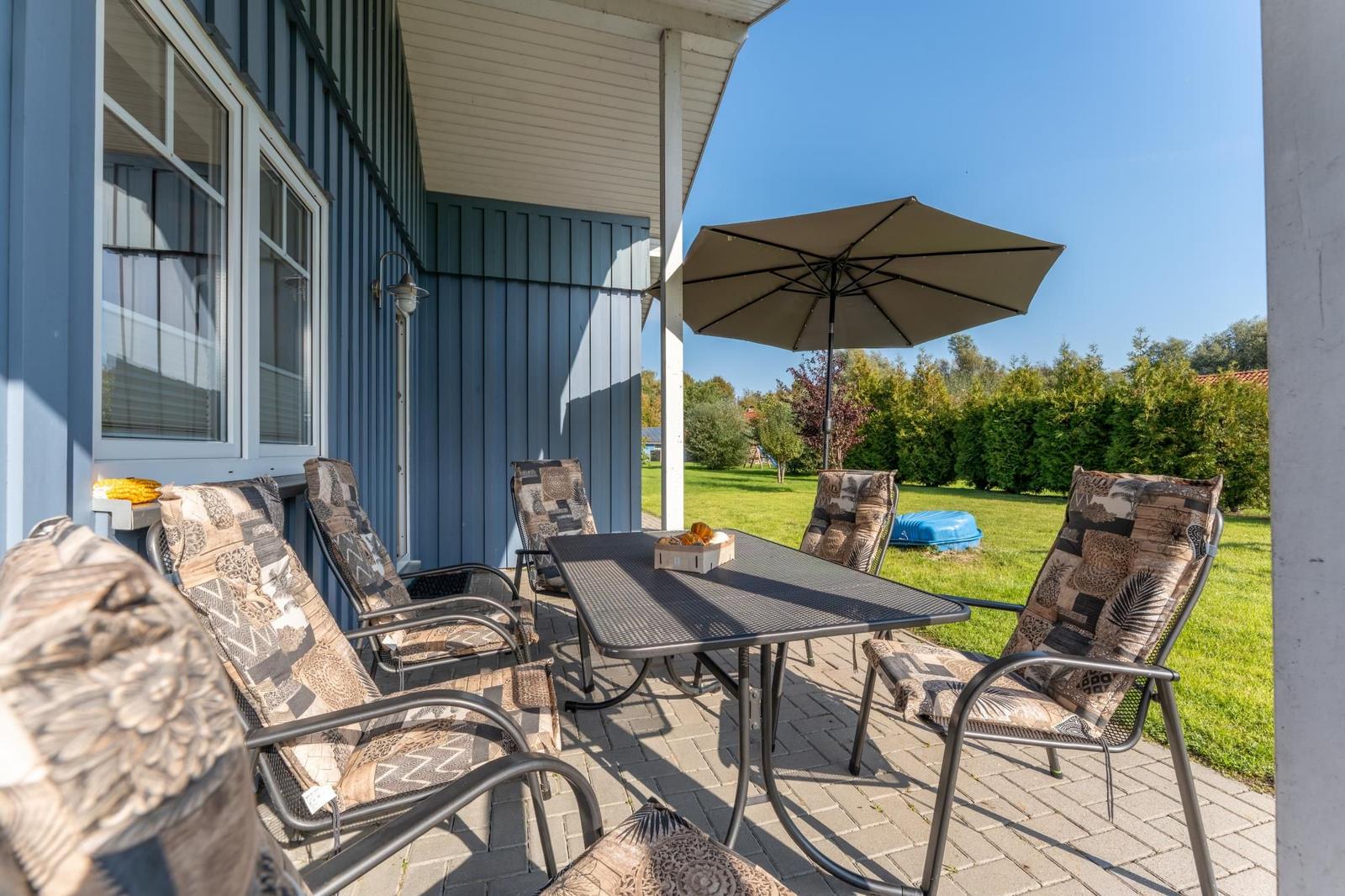 Terrasse mit Tisch, Stühlen und Sonnenschutz unter blauem Himmel.
