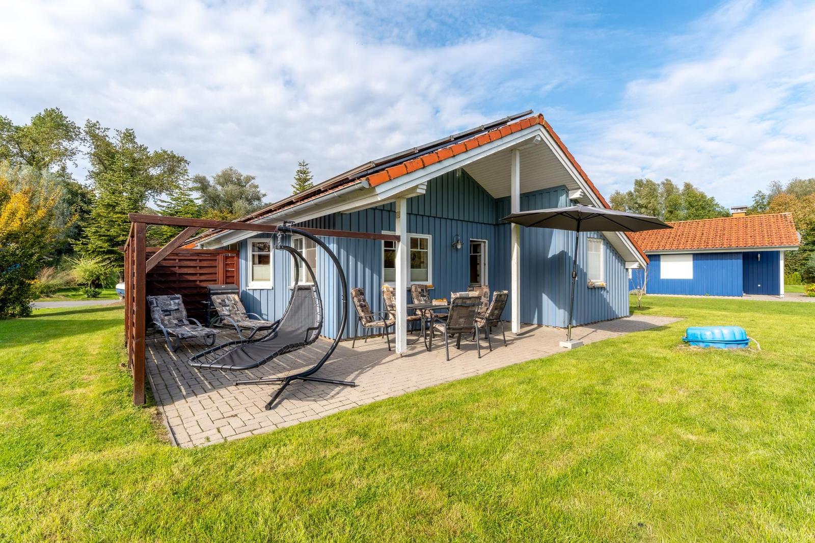 Blauer Ferienhaus mit Terrasse, Liege und Sitzgruppe im Garten.
