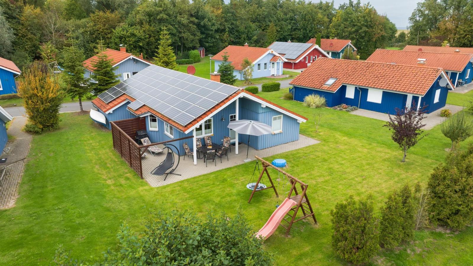 Blauer Ferienhaus mit Solaranlage, Terrasse und Spielplatz auf grünem Grund.