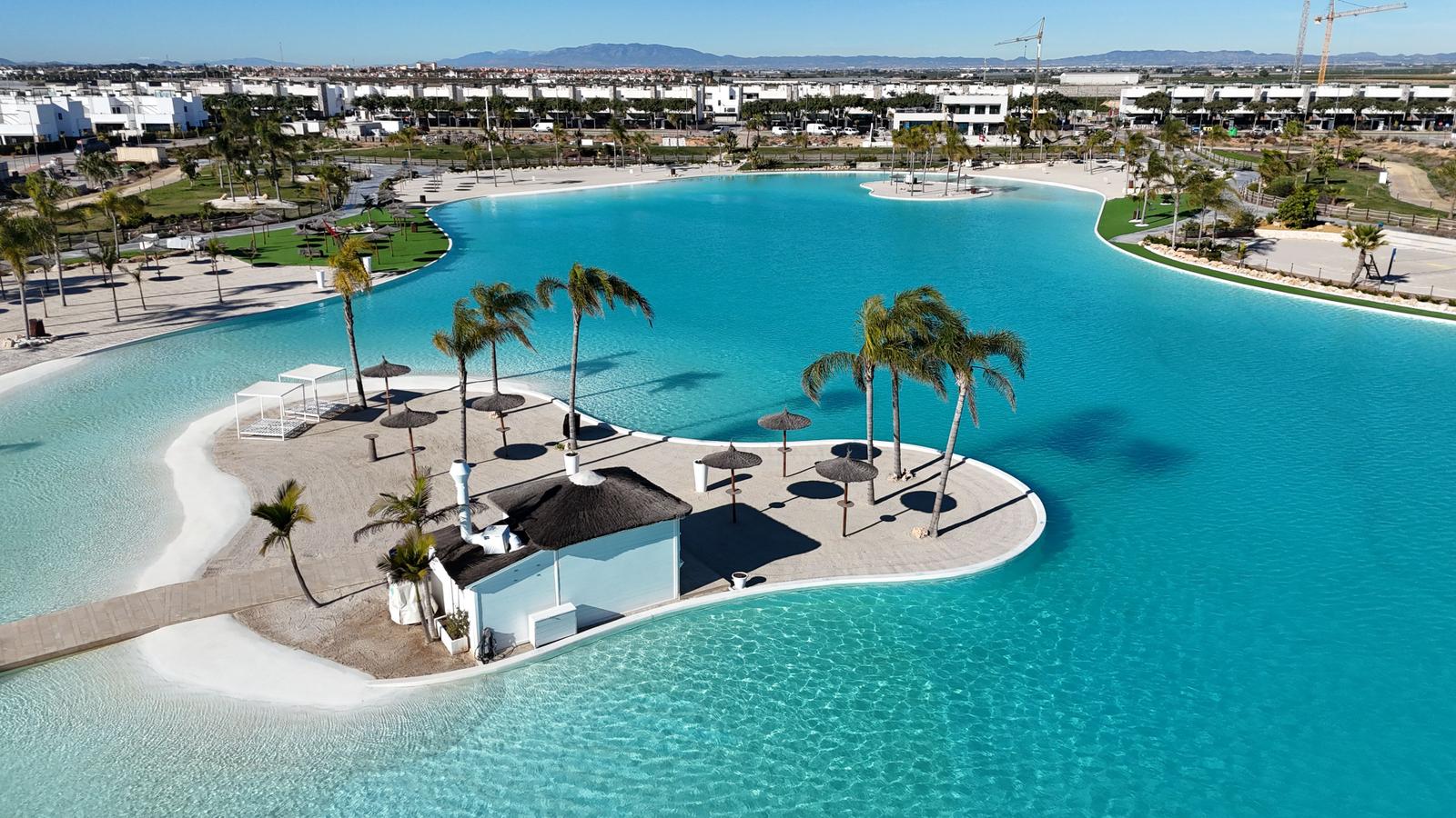 Large pool with island, palms, and umbrellas. Background: residential area and mountains.