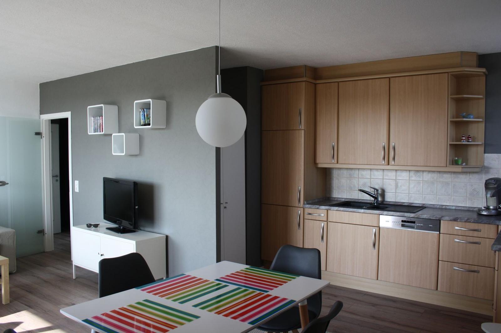 Küche mit Holzschränken, Essbereich mit Tisch und Stühlen, Fernseher und Wandregale.