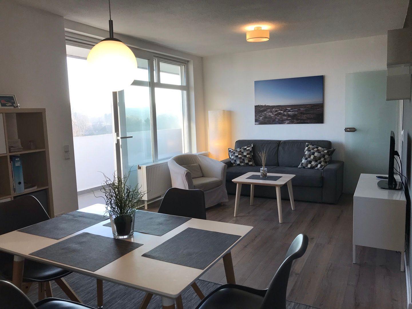 Wohn- und Essbereich mit Sofa, Tisch und Stühlen. Große Fenster und Holzboden.