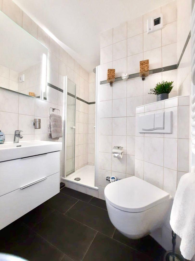 Badezimmer mit Dusche, Waschbecken und Toilette. Weiße Fliesen und dunkler Boden.