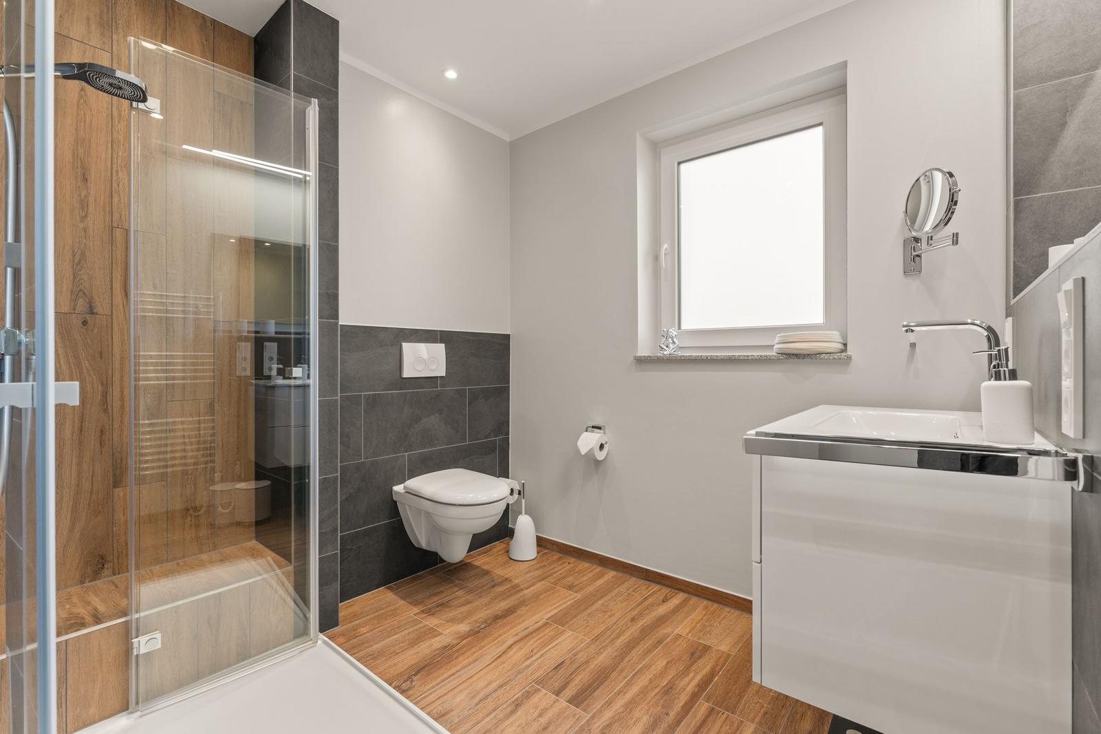 Badezimmer mit Dusche, Toilette und Waschbecken. Holzboden und Fenster.