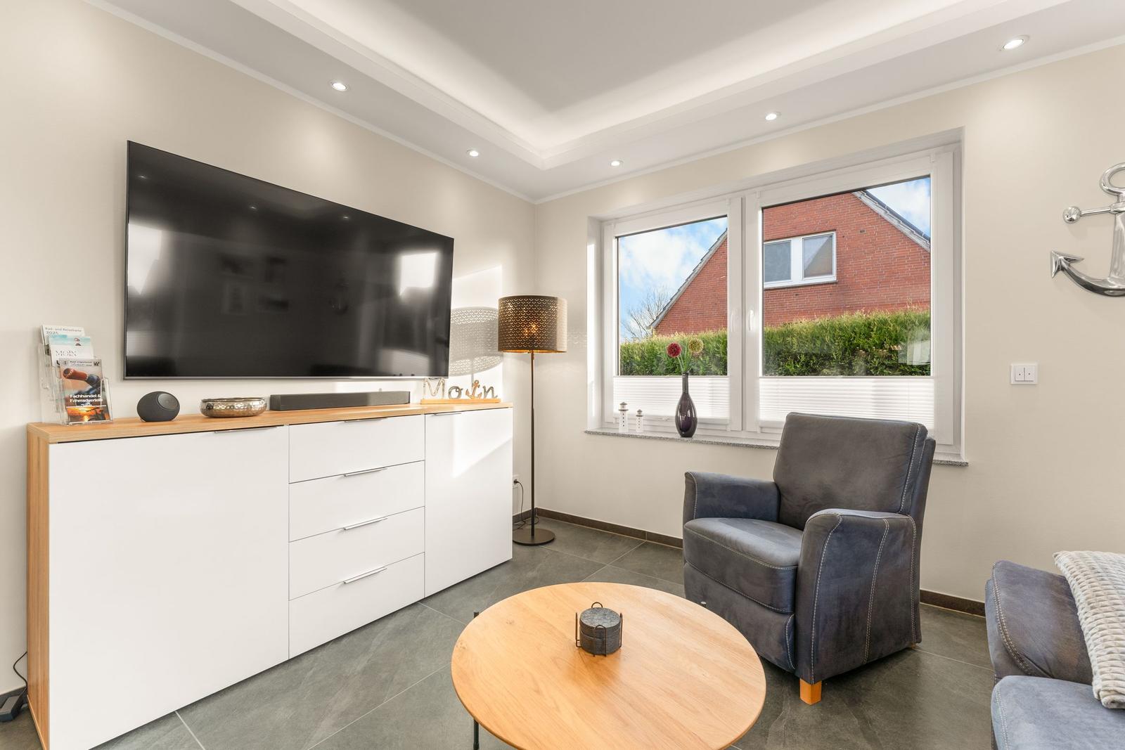 Wohnzimmer mit TV, Sessel und Tisch. Fenster zeigt Gartenansicht.