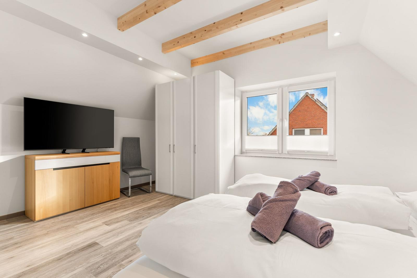 Schlafzimmer mit Doppelbett, TV, Kleiderschrank und Fensterblick auf Haus.