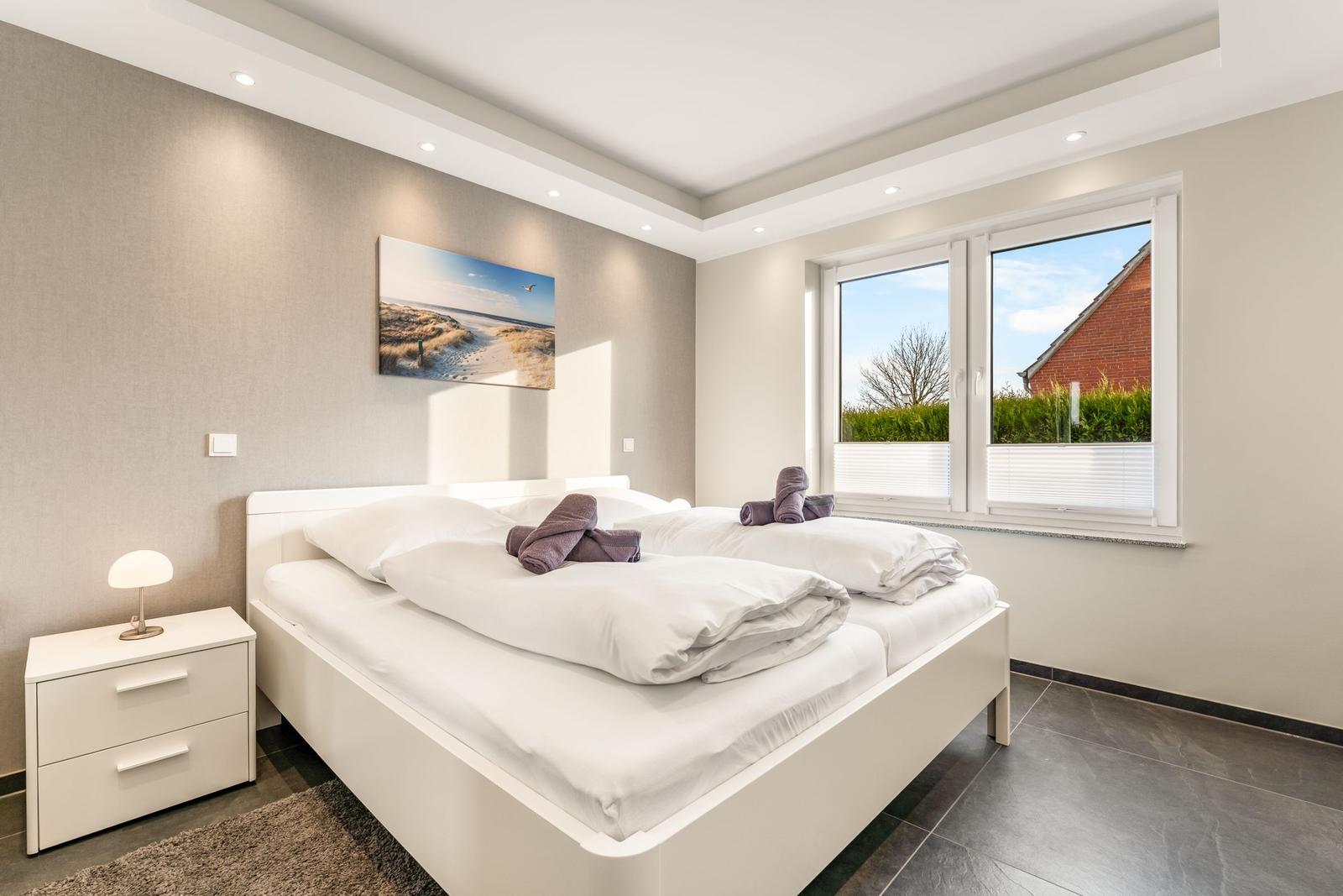 Schlafzimmer mit weißem Bett, Nachttisch und Fenster mit Blick auf Garten.