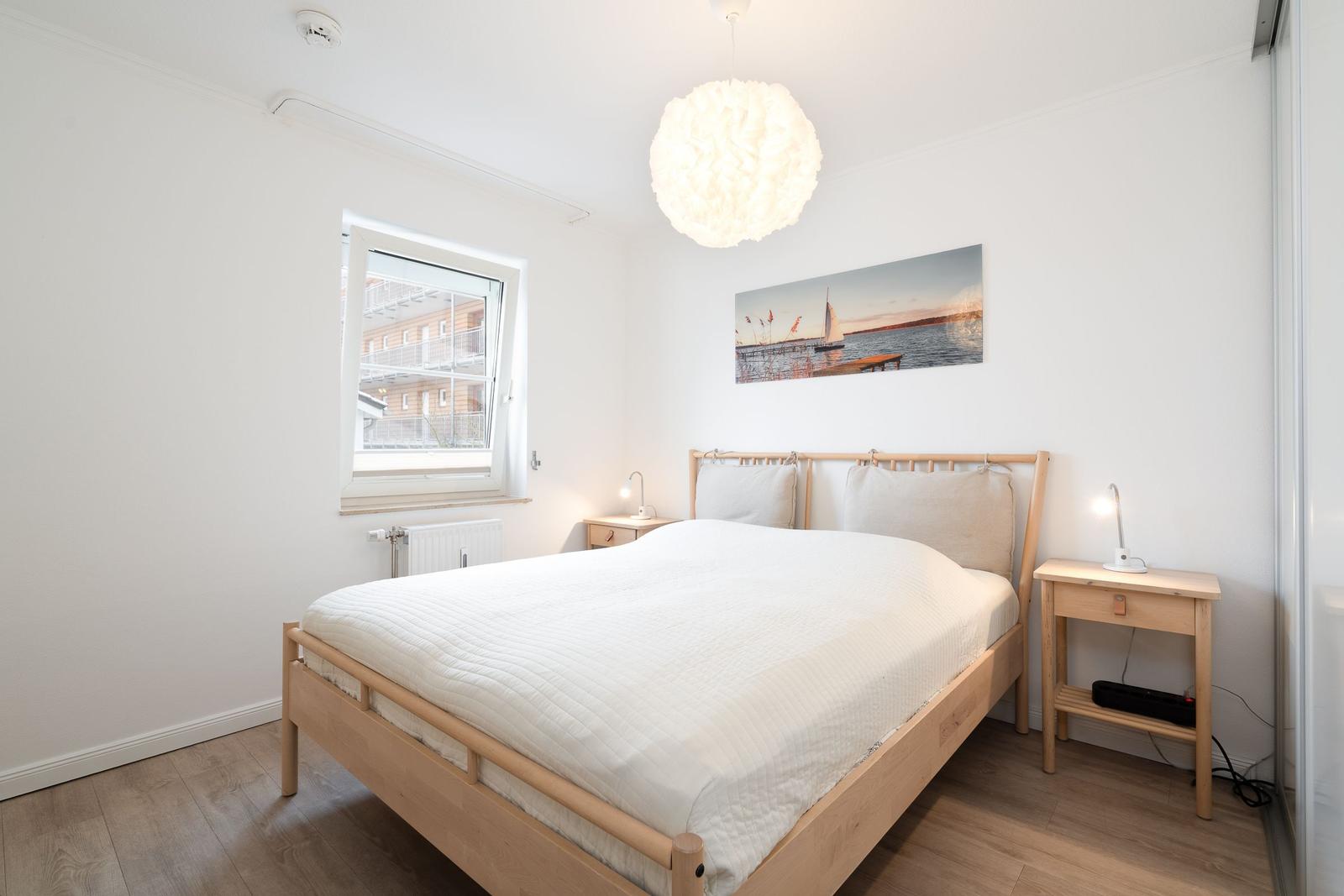 Schlafzimmer mit Holzbett, Nachttischen und Wandbild. Fenster mit Blick auf Gebäude.