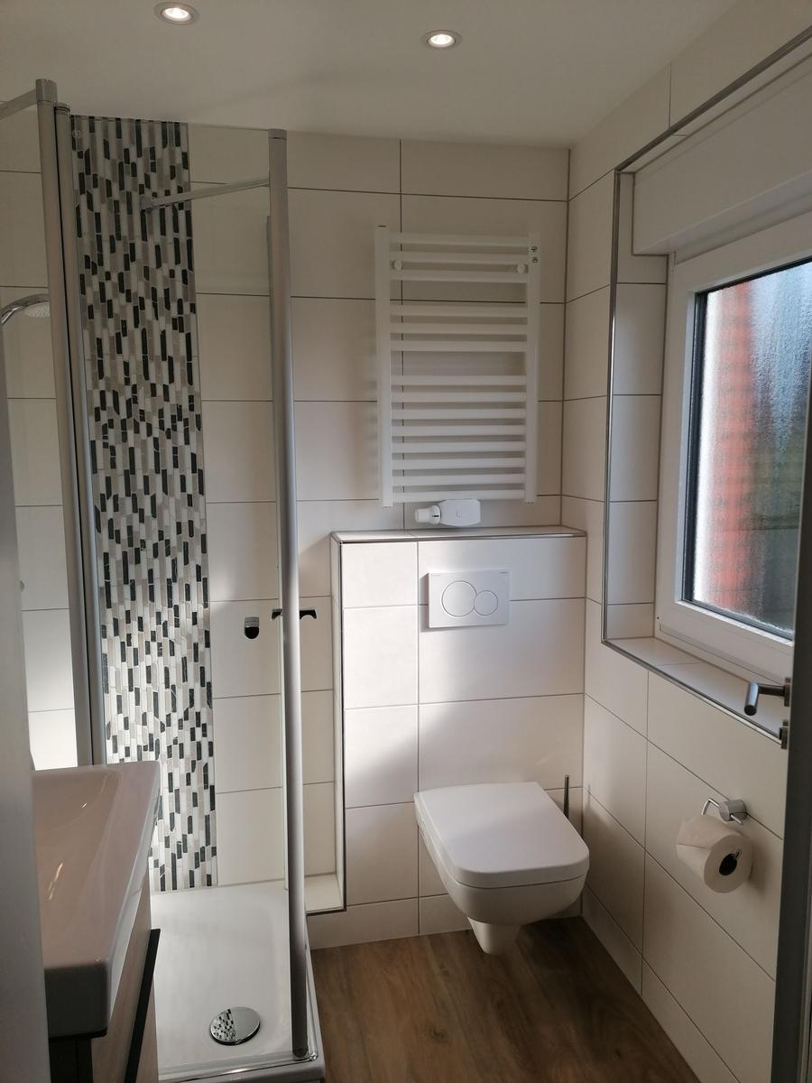 Badezimmer mit Dusche, Toilette und Fenster. Weiße Fliesen und Holzfußboden.