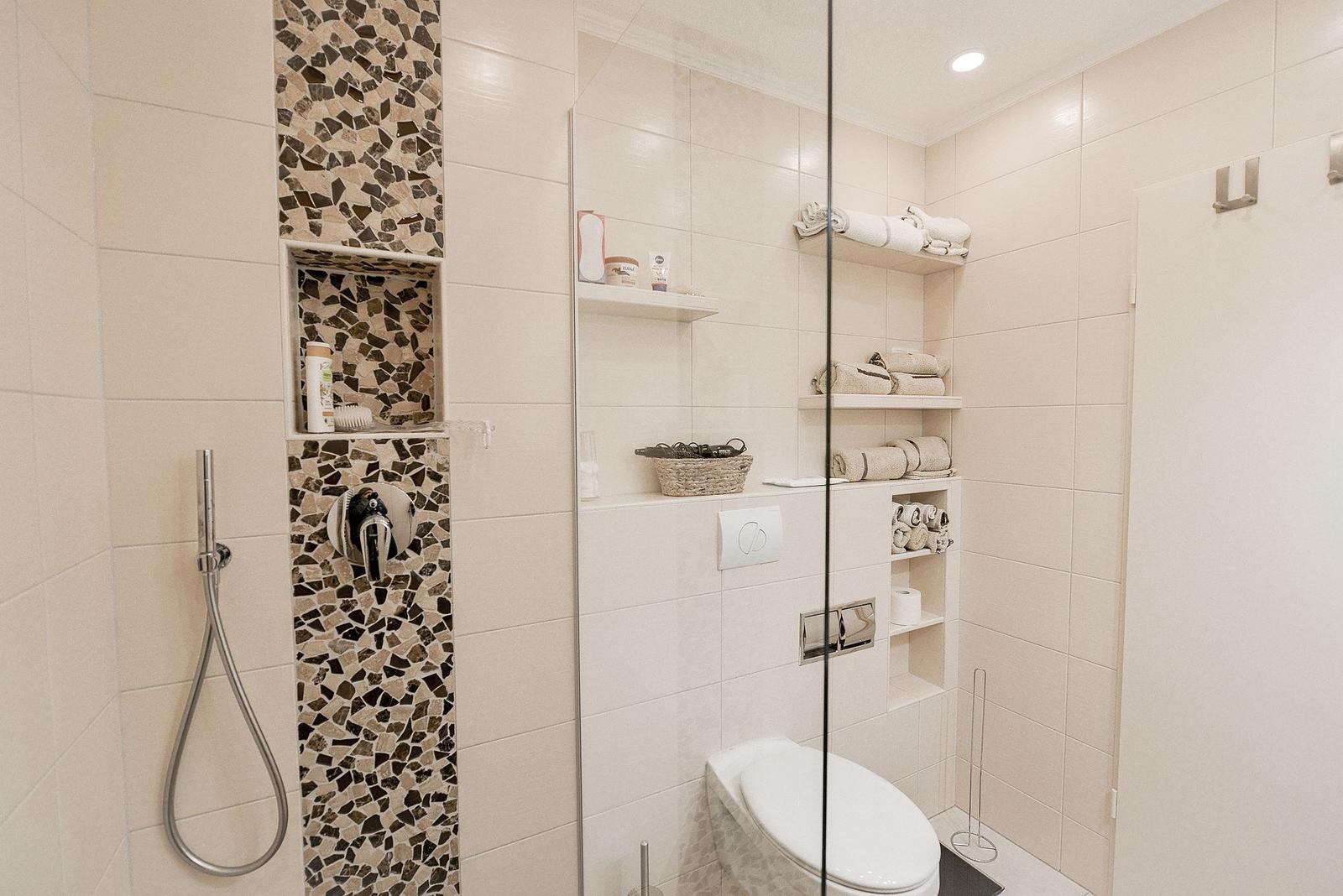Dusche mit Mosaikwand, Toilette und integrierte Regale mit Handtüchern.