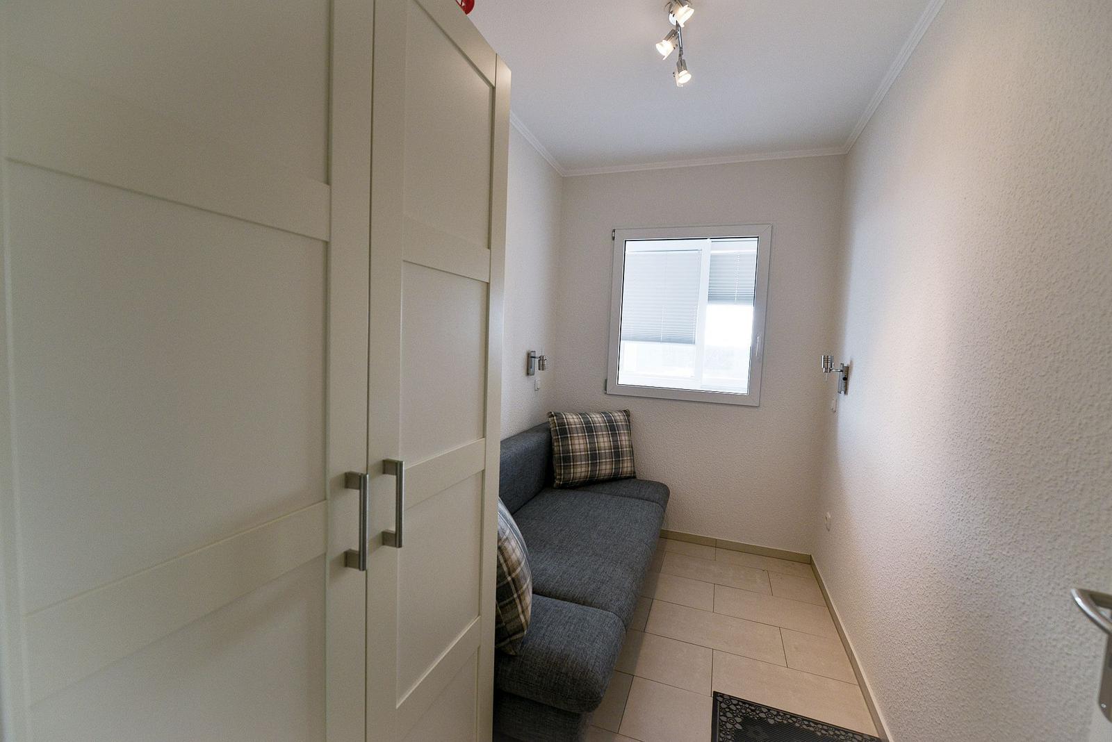 Ein kleiner Raum mit grauem Sofa, weißen Wänden und einem Fenster mit Jalousie.