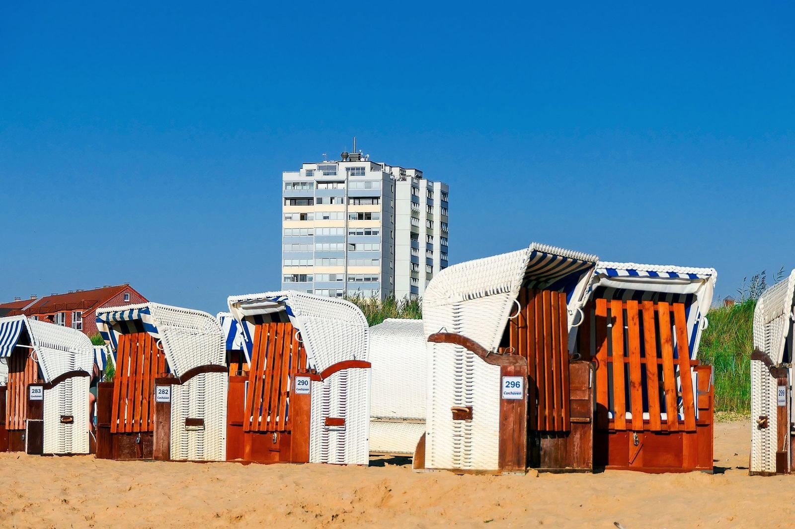 Strandkörbe auf Sandstrand mit Hochhaus im Hintergrund.