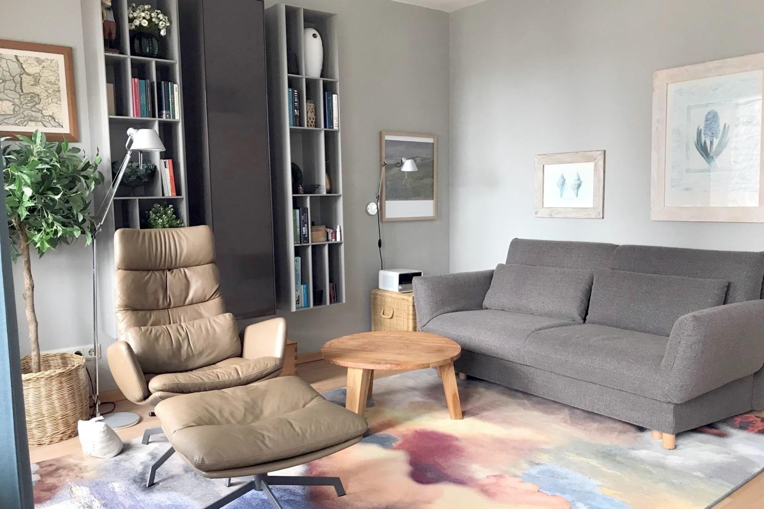 Wohnzimmer mit grauem Sofa, braunem Sessel und runden Tisch auf buntem Teppich.