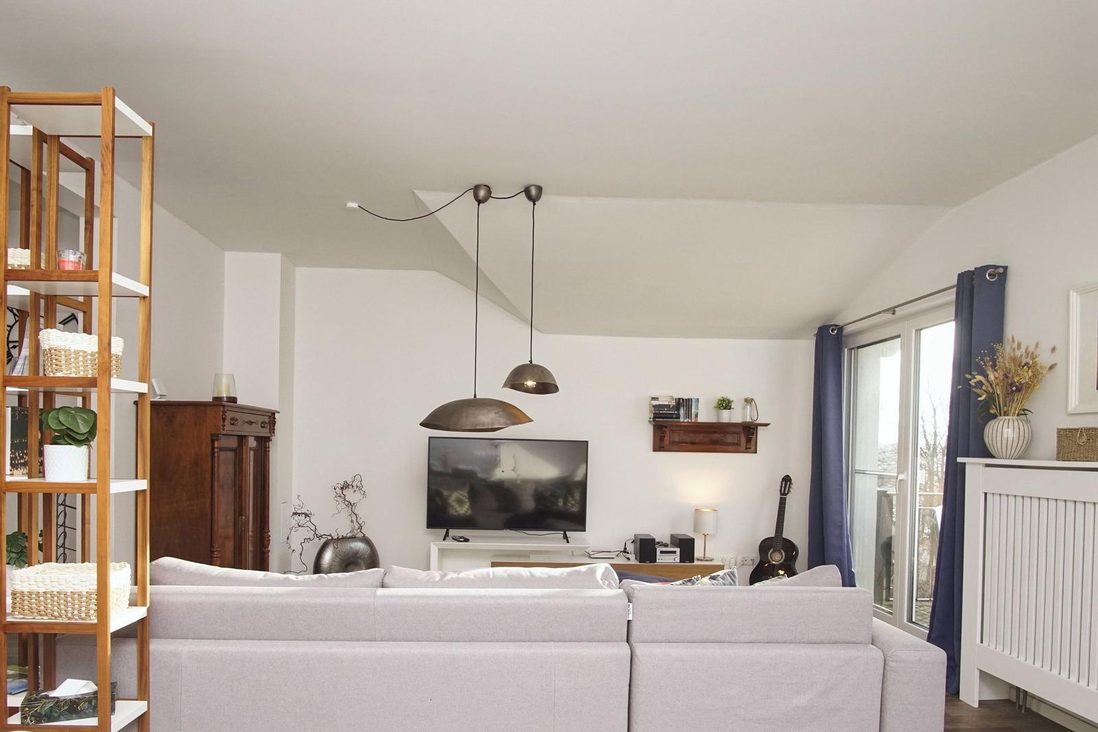 Wohnzimmer mit Sofa, Fernseher und Schwebelampen. Fenster mit blauen Vorhängen.