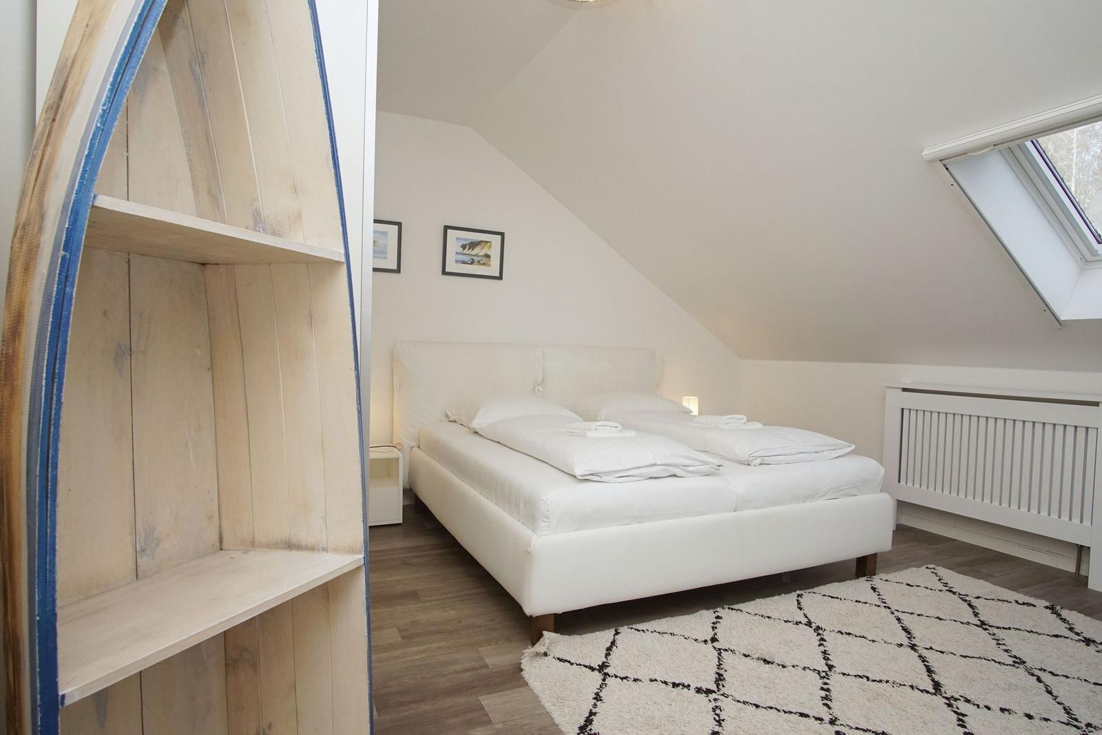 Schlafzimmer mit Dachschrägen, weißem Bett und Dachfenster. Holzregal im Vordergrund.