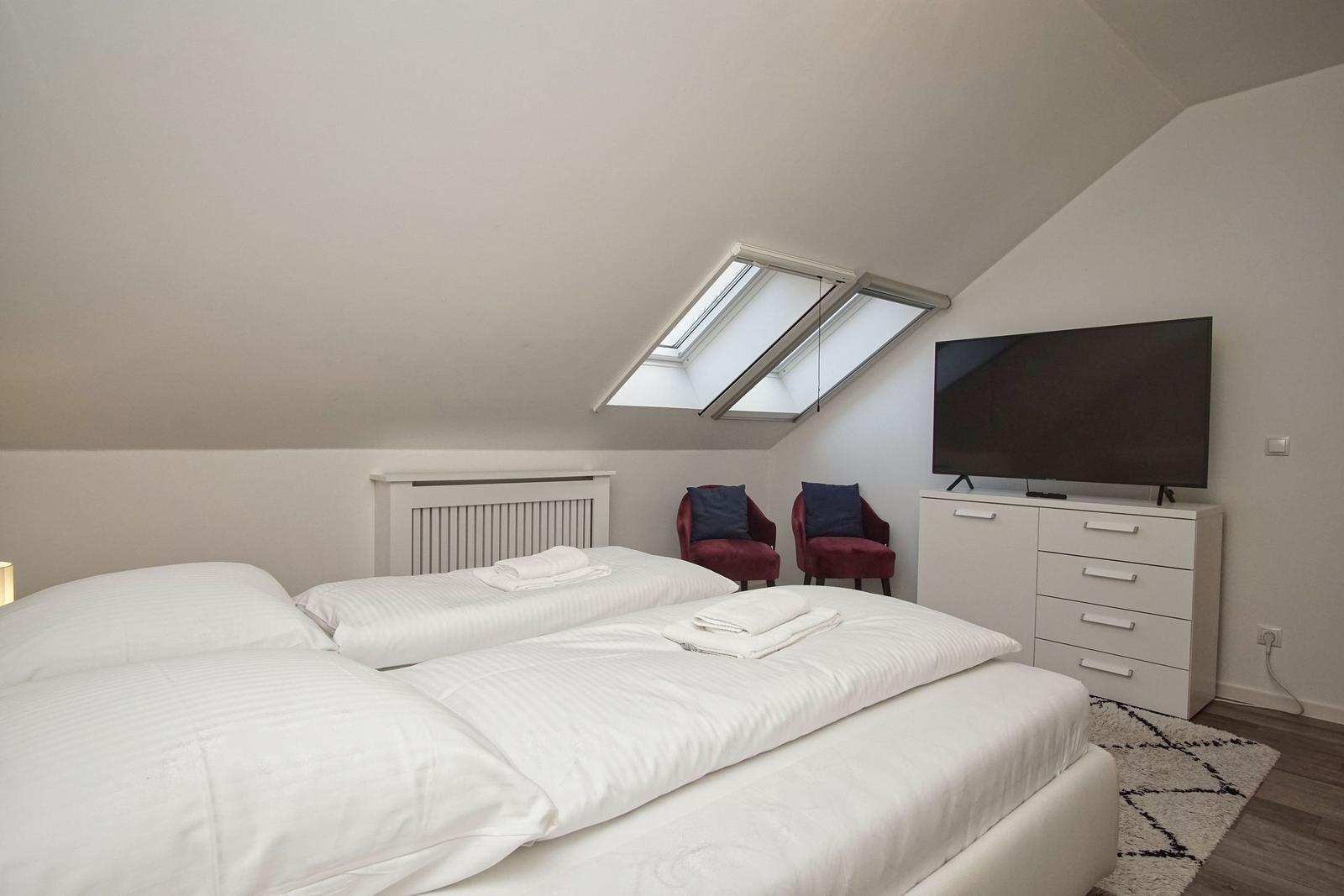 Doppelbett mit weißer Bettwäsche, zwei rote Sessel, TV und Dachfenster.