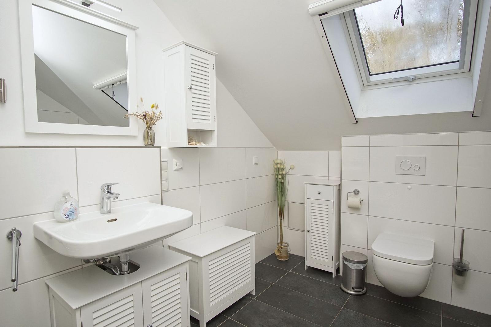 Badezimmer mit Dusche, Spiegel und Fenster. Weiße Fliesen und dunkle Bodenfliesen.