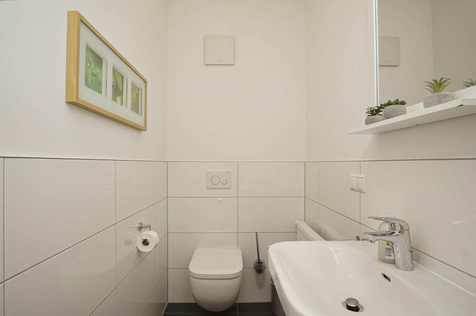 Badezimmer mit Toilette, Waschbecken und Spiegel. An der Wand hängt ein Bild mit Pflanzenmotiv.