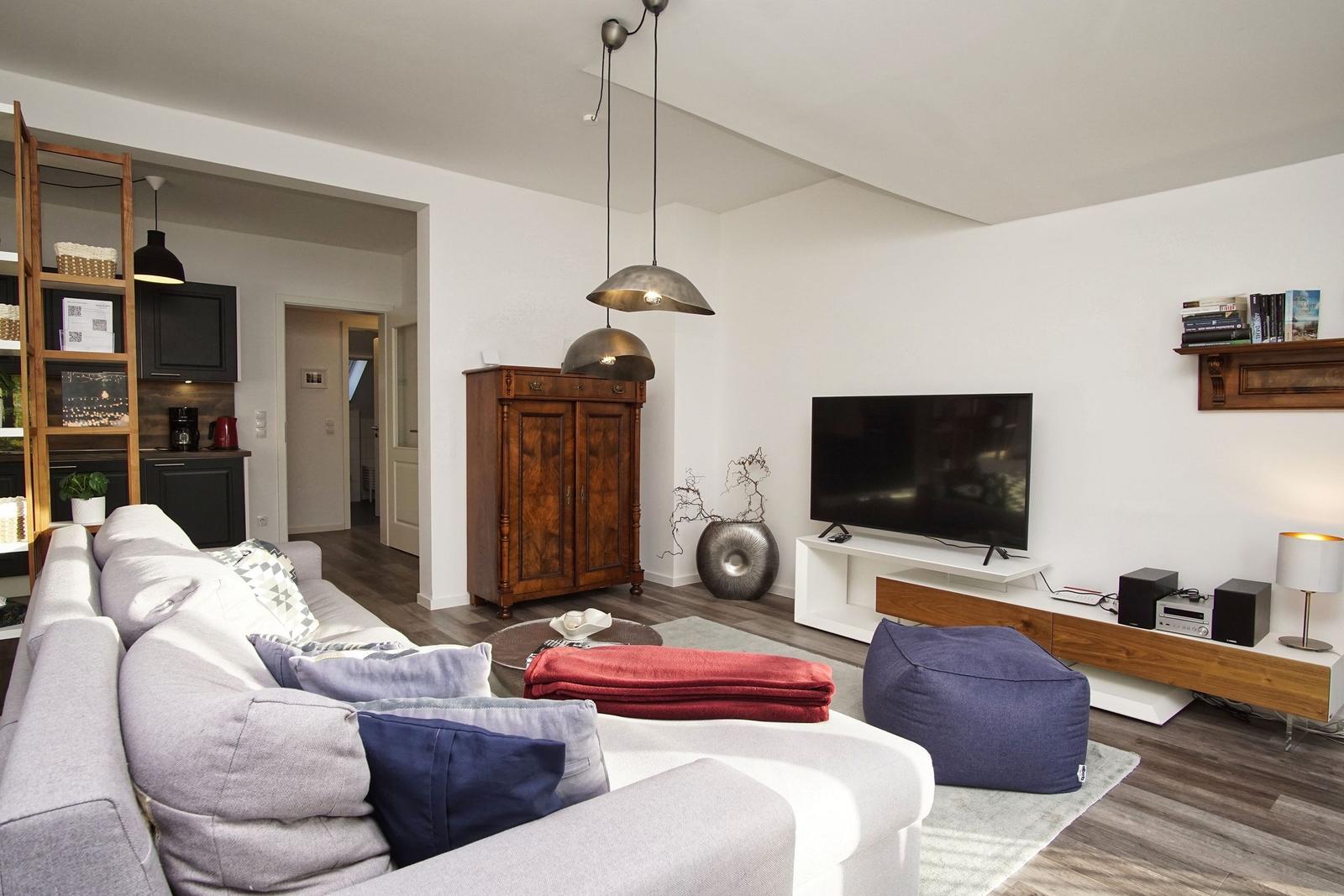 Wohnzimmer mit Sofa, Fernseher und Holzmöbel. Kühlschrank und Küchenzeile im Hintergrund.