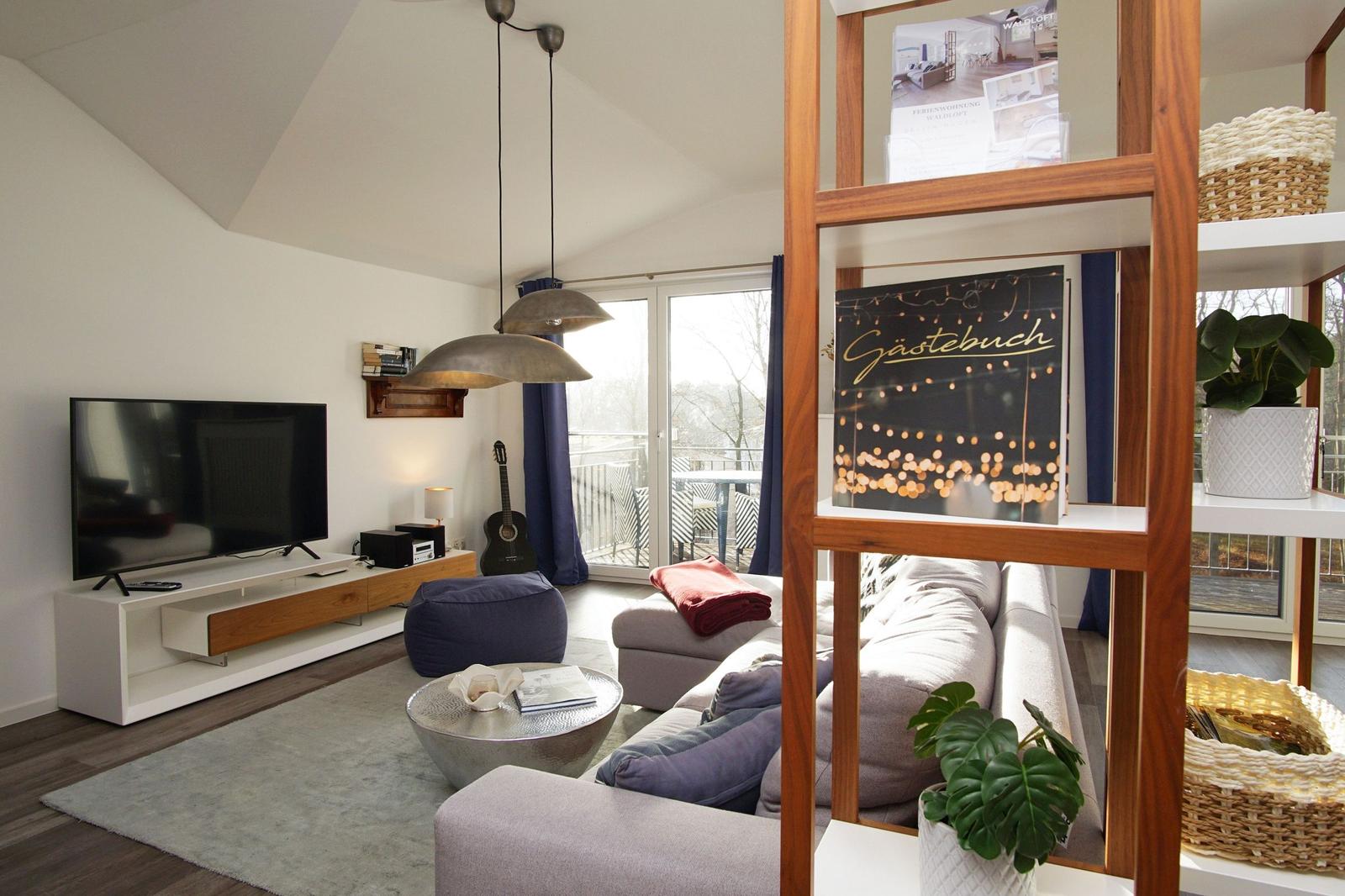 Wohnzimmer mit Sofa, Fernseher und Terrasse. Holzregal, Pflanzen und Sitzkissen.