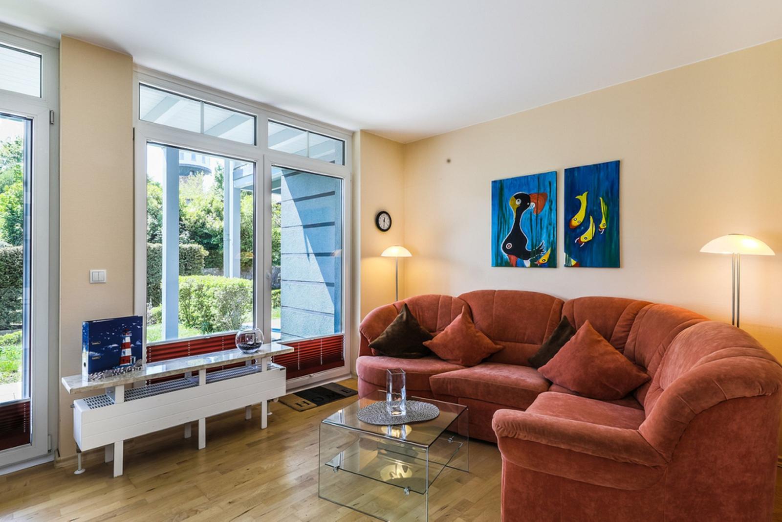 Wohnzimmer mit rotem Sofa, Glas-Tisch und großen Fenstern. Wandbilder zeigen Fische.