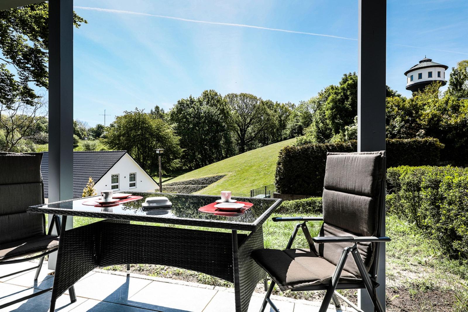 Terrasse mit Tisch und Stühlen. Blick auf grüne Hügel und einen Wasserturm.
