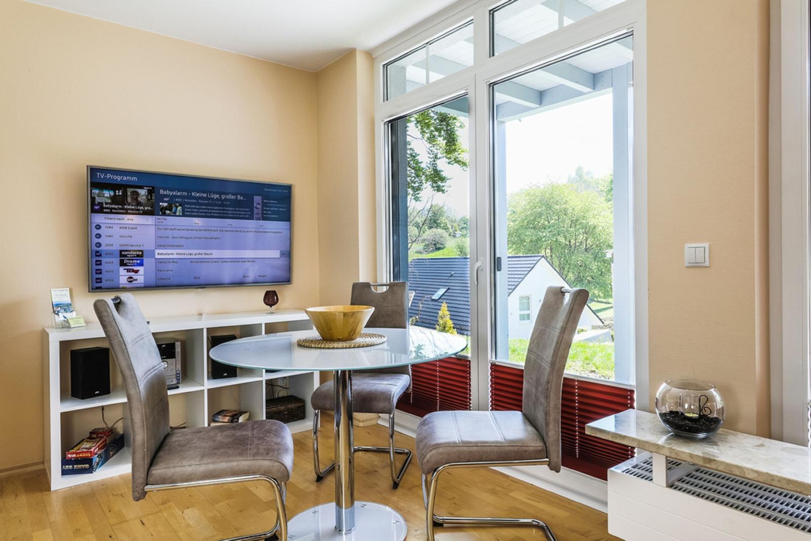 Essbereich mit Glasrundtisch, Stühlen und TV. Blick auf Garten durch große Fenster.