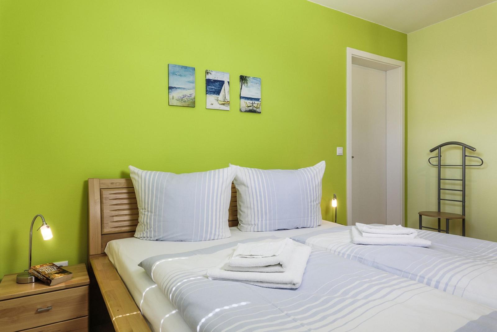 Schlafzimmer mit grüner Wand, Bett mit Bettwäsche und Handtüchern, Nachttisch und Bildern.