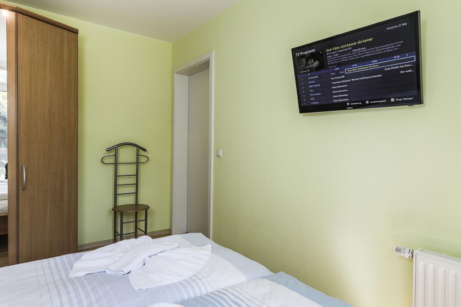 Schlafzimmer mit Bett, Kleiderständer, Schrank und Wandfernseher.