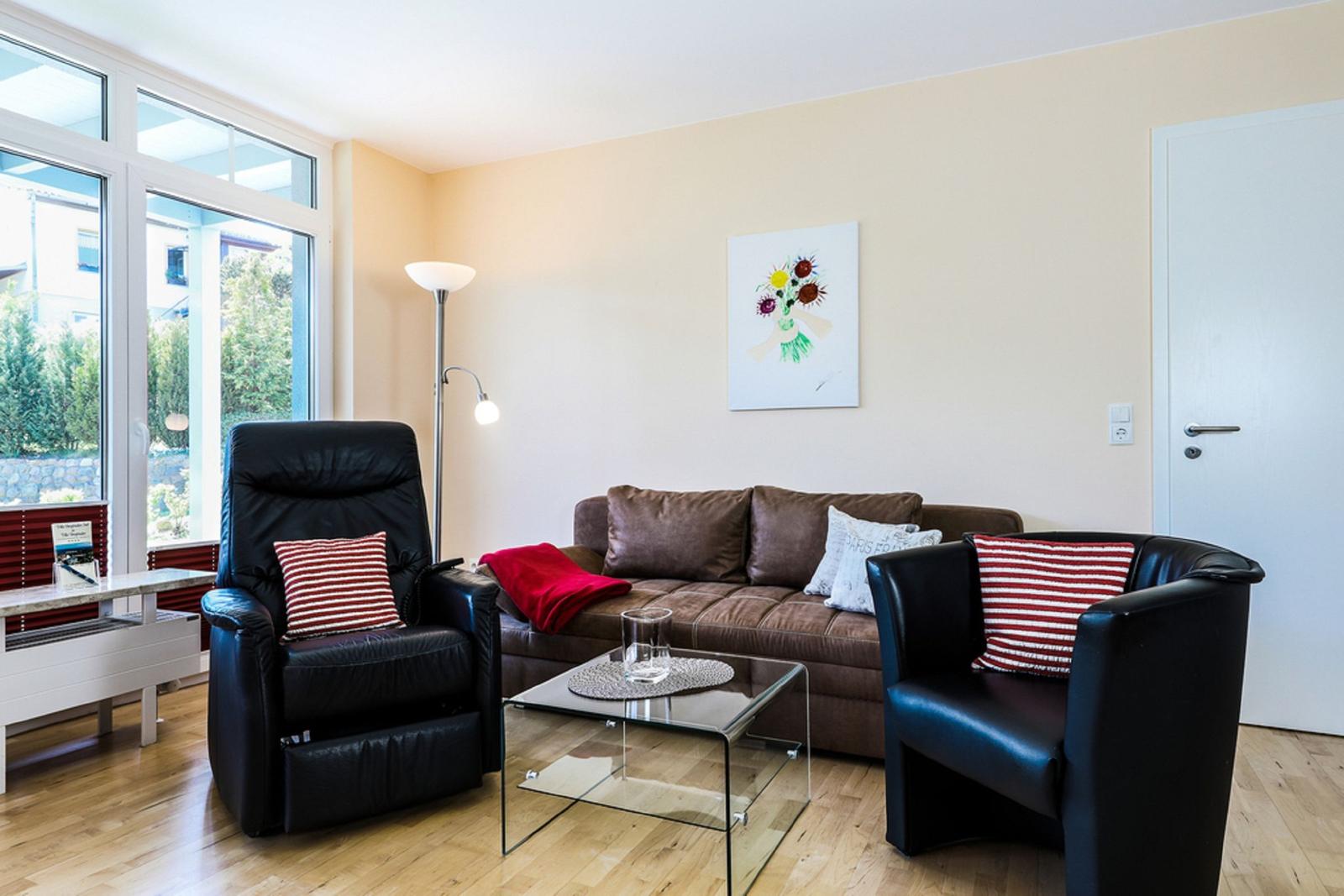 Wohnzimmer mit braunem Sofa, zwei schwarzen Sesseln und Glas-Tisch. Große Fenster mit Blick nach draußen.